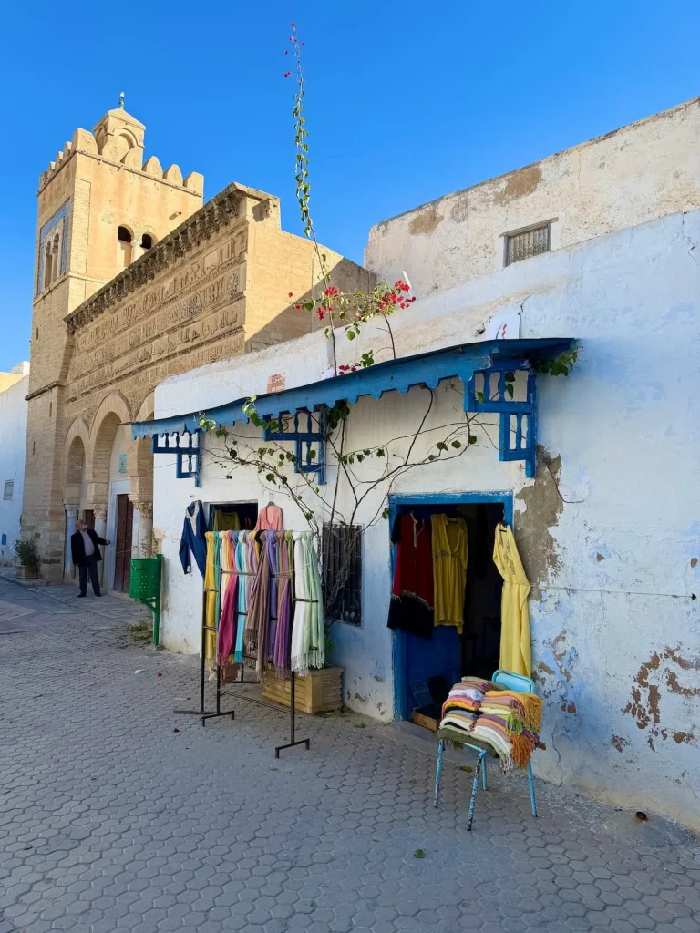 Kairouan Medina with a clothes stall selling shawls next door to the 3 doors mosque with a man standing outside, Tunisia. The walls of teh Medina are white washed with writing on the walls.
