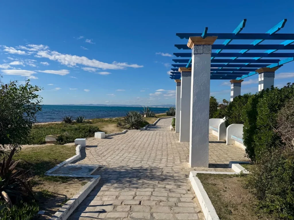 Promenade with sea to the left, foliage along the sandy path and seating area to the right in Hammamet
