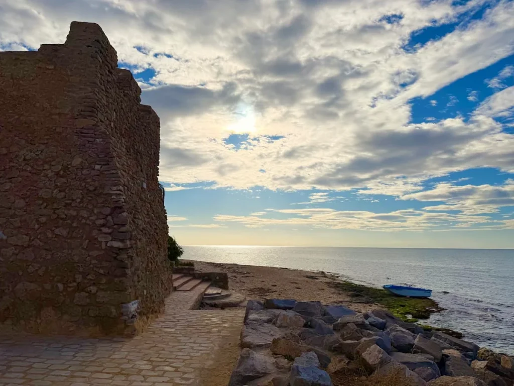 Side of the Kasbah Fort with a fishing boat and sea in Hammamet. There are some defence rocks on the path along the sand